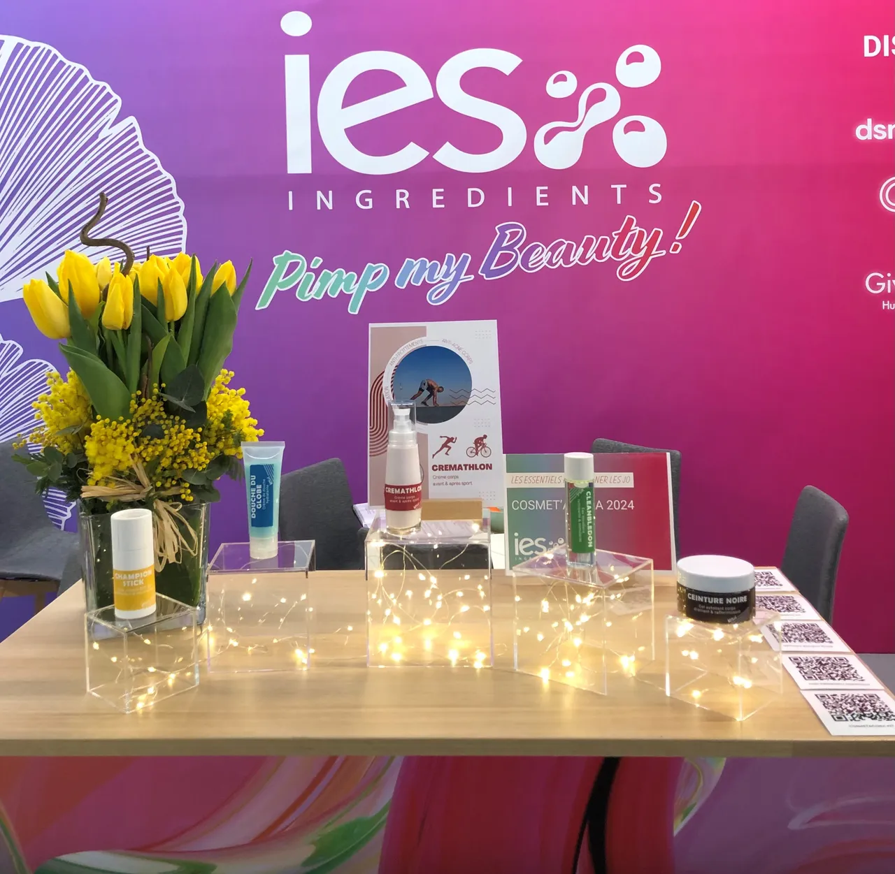 Stand d'exposition IES Ingredients avec produits cosmétiques, fleurs jaunes et logo Pimp my Beauty sur fond rose-violet