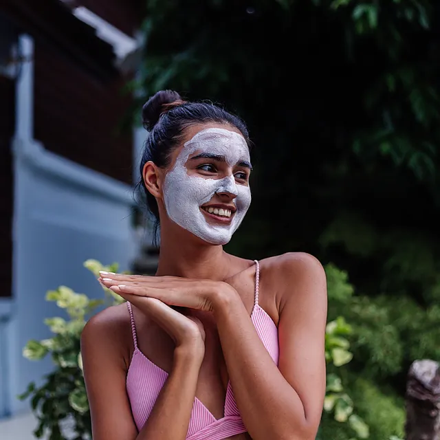 Smiling woman applying a white facial mask outdoors, natural cosmetics